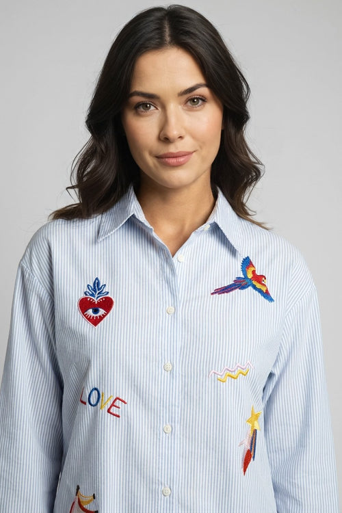 Modelo con camisa de rayas azul y bordados coloridos, vista frontal detalle pecho con loro y corazón bordados