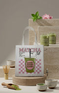 Tote bag blanca con ilustracion retro de iced matcha latte sobre cuadros rosas, vista frontal